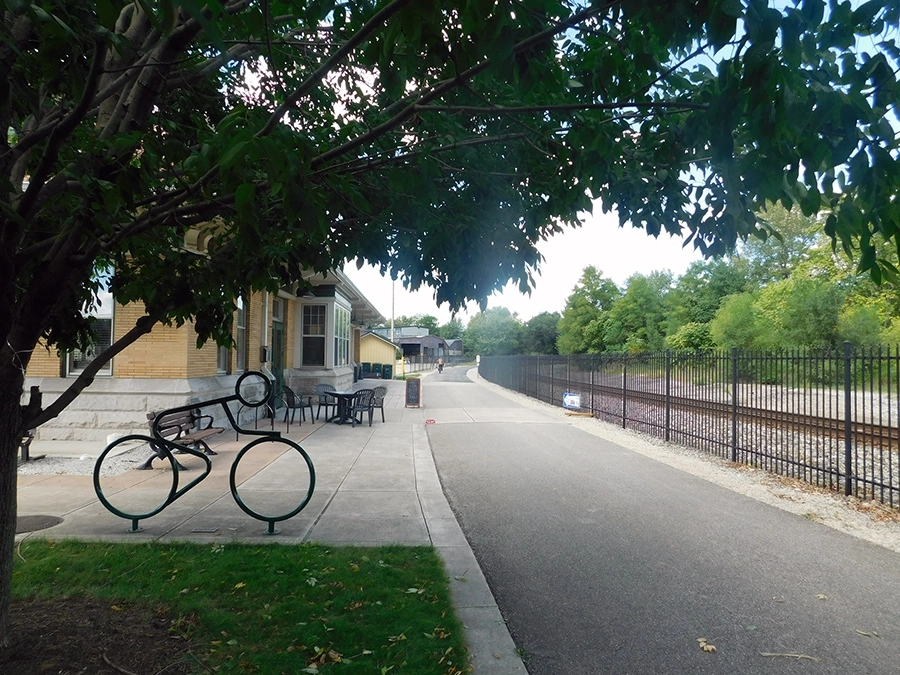A bicycle staple shaped like a person on a bike next to a paved trail and tan brick building.