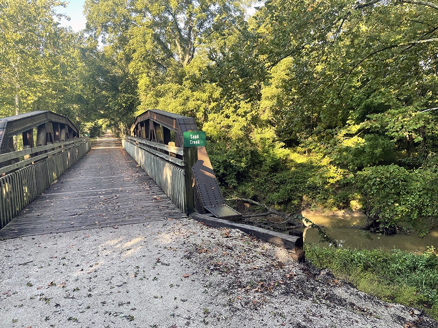 A sign on a wooden bridge says "Sand Creek" with a muddy creek passing under.