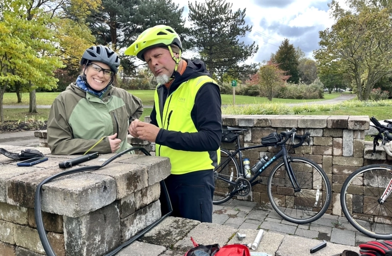 two cyclists smile at the camera, they look like they are changing a flat tire