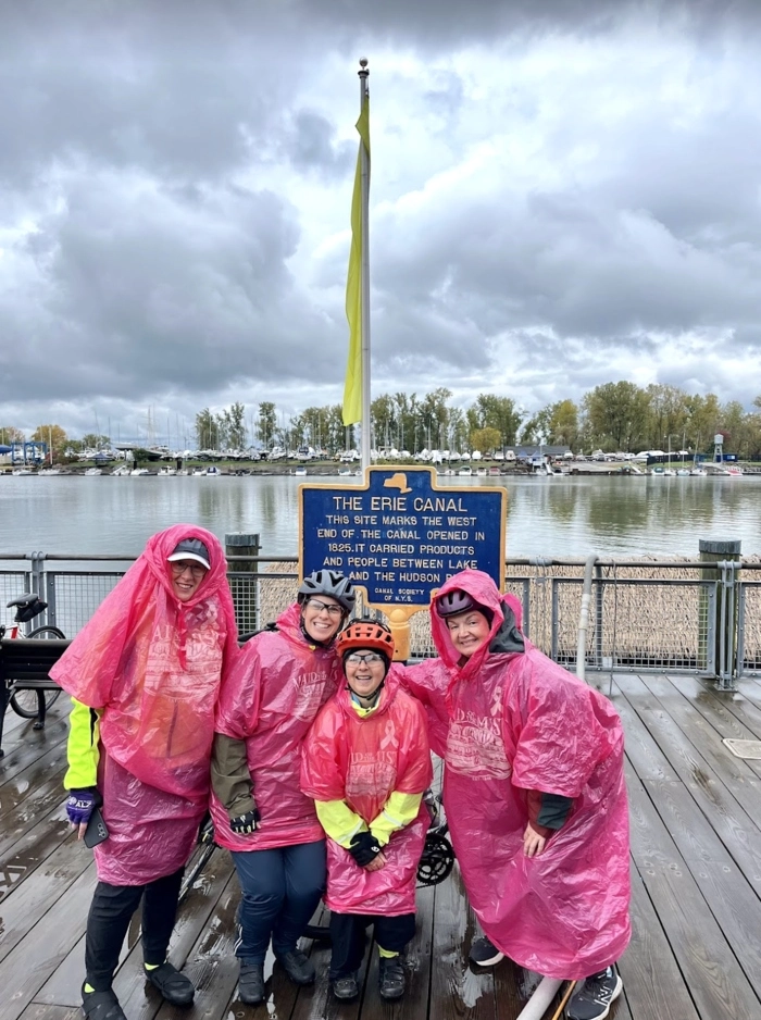 four cyclists in pink ponchos stand in front of a canal trail sign with a river and cloudy sky in the background