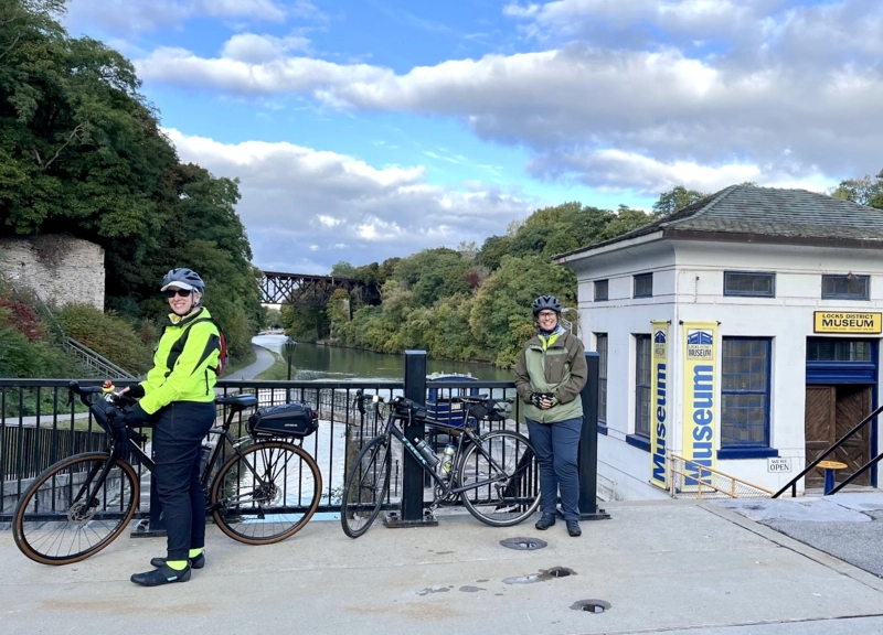 two cyclists stand on a bridge over a river with trees and a blue sky in the background