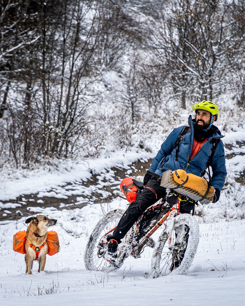 In a snow winter landscape, a man straddles his bike and his dog watches him intently