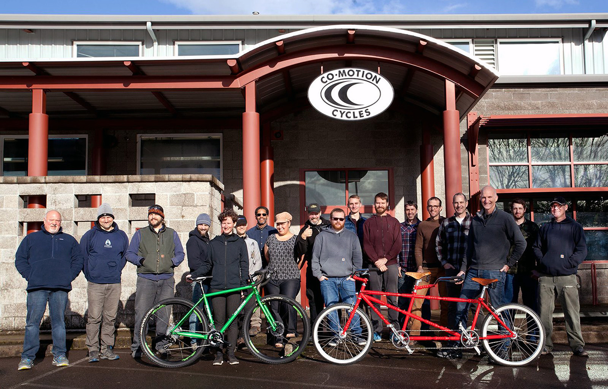 A large group of people stand outside of Co-Motion Cycles headquarters, a large warehouse.