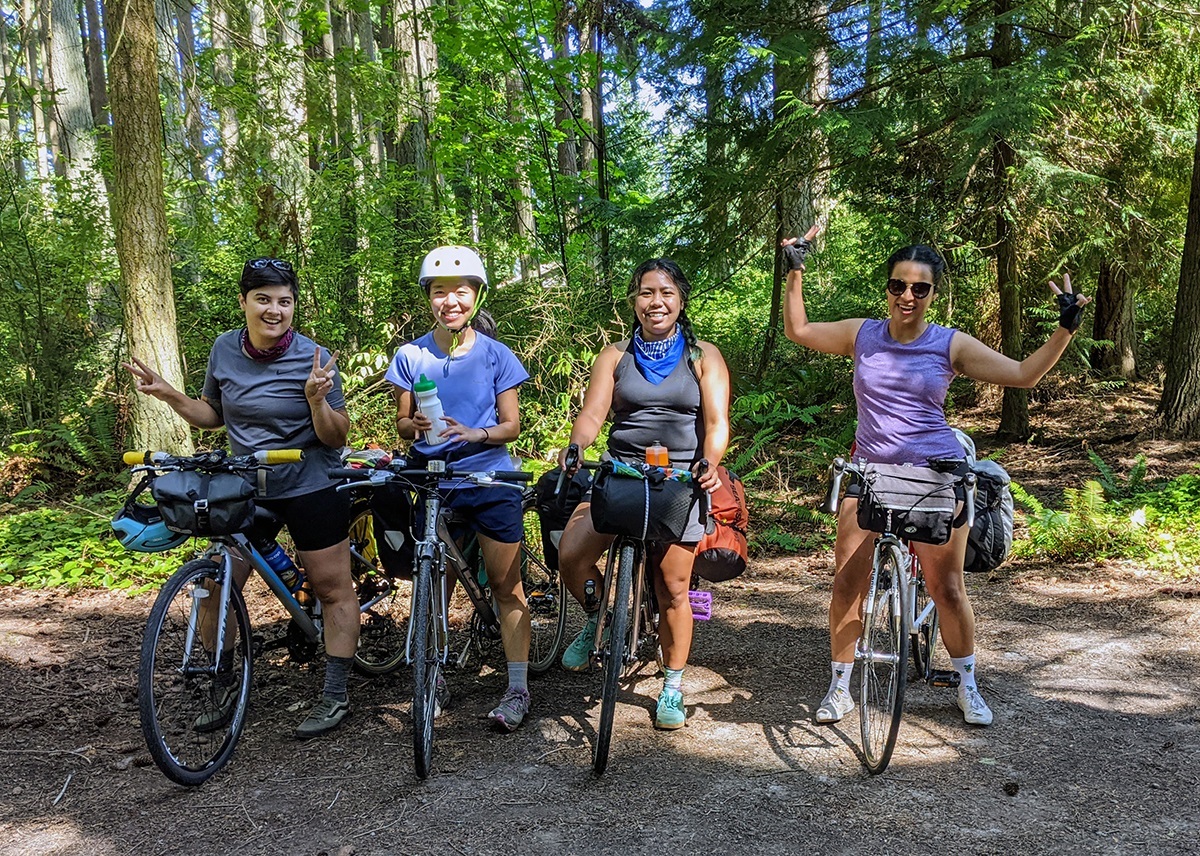 Newbie bikecampers pose for a photo among the tall trees of Kitsap Memorial Park. Each rider is straddling their loaded bikes with various poses and all smiling. The speckled sunshine beams upon their bikes and bodies.