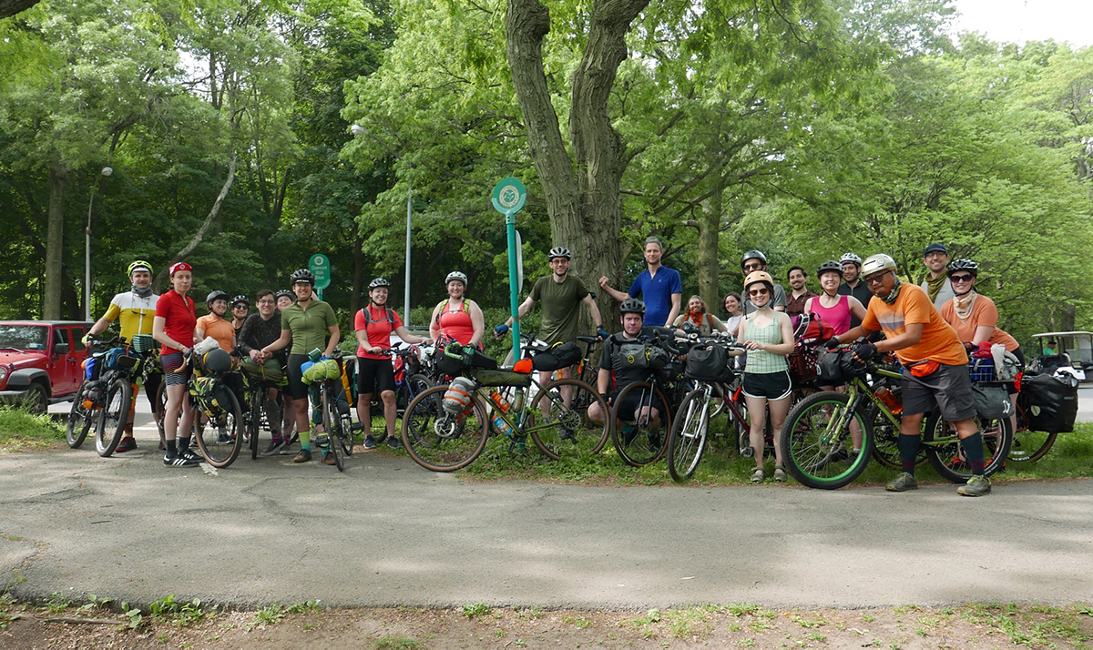 A large group gathers together with their loaded bikes under the shade trees to snap a photo
