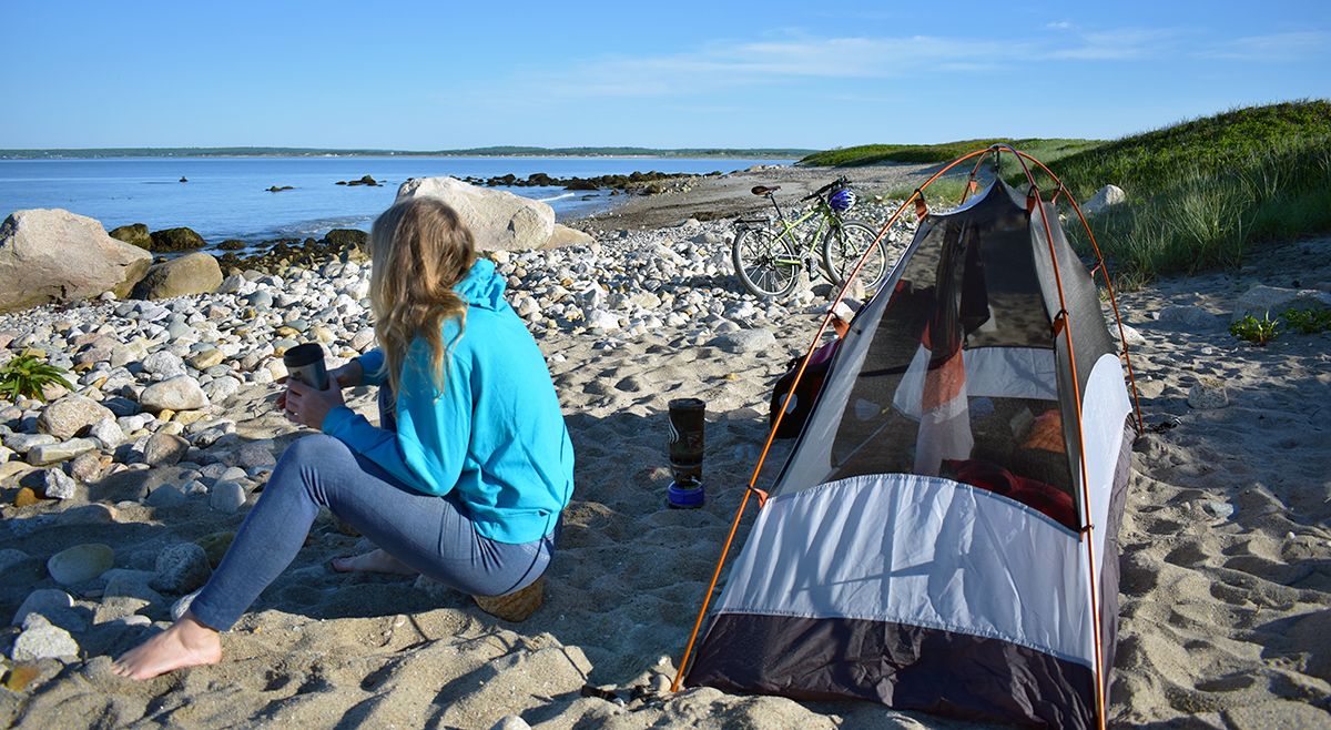 Laura sits with a hot cup of coffee in front of her tent on the ocean shore.