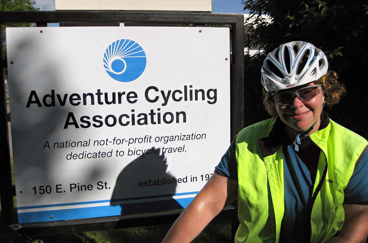 Donna stands in her cycling gear next to the Adventure Cycling headquarters sign in Missoula, Montana