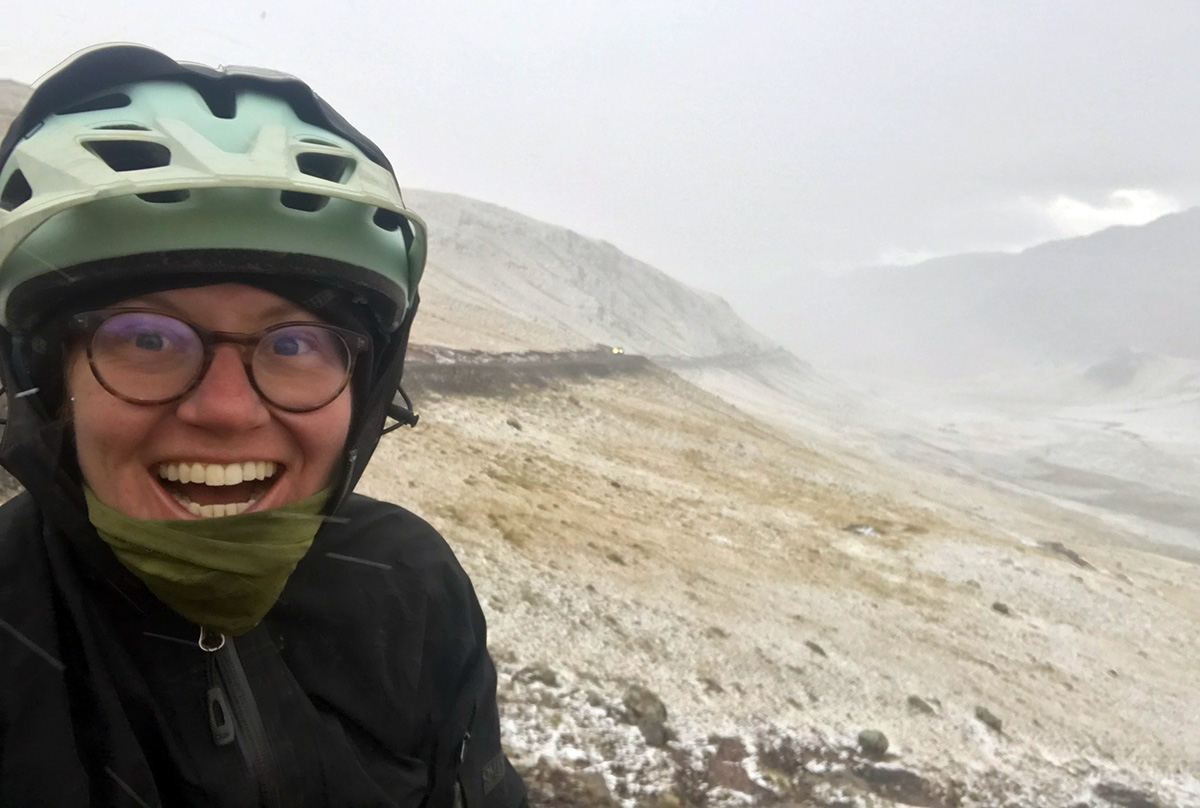 Hollie takes a selfie on top of a mountain pass. Visibility is low due to snow and hail and low clouds but Hollie has a huge smile on her face as she's wrapped up tight in lots of layers.