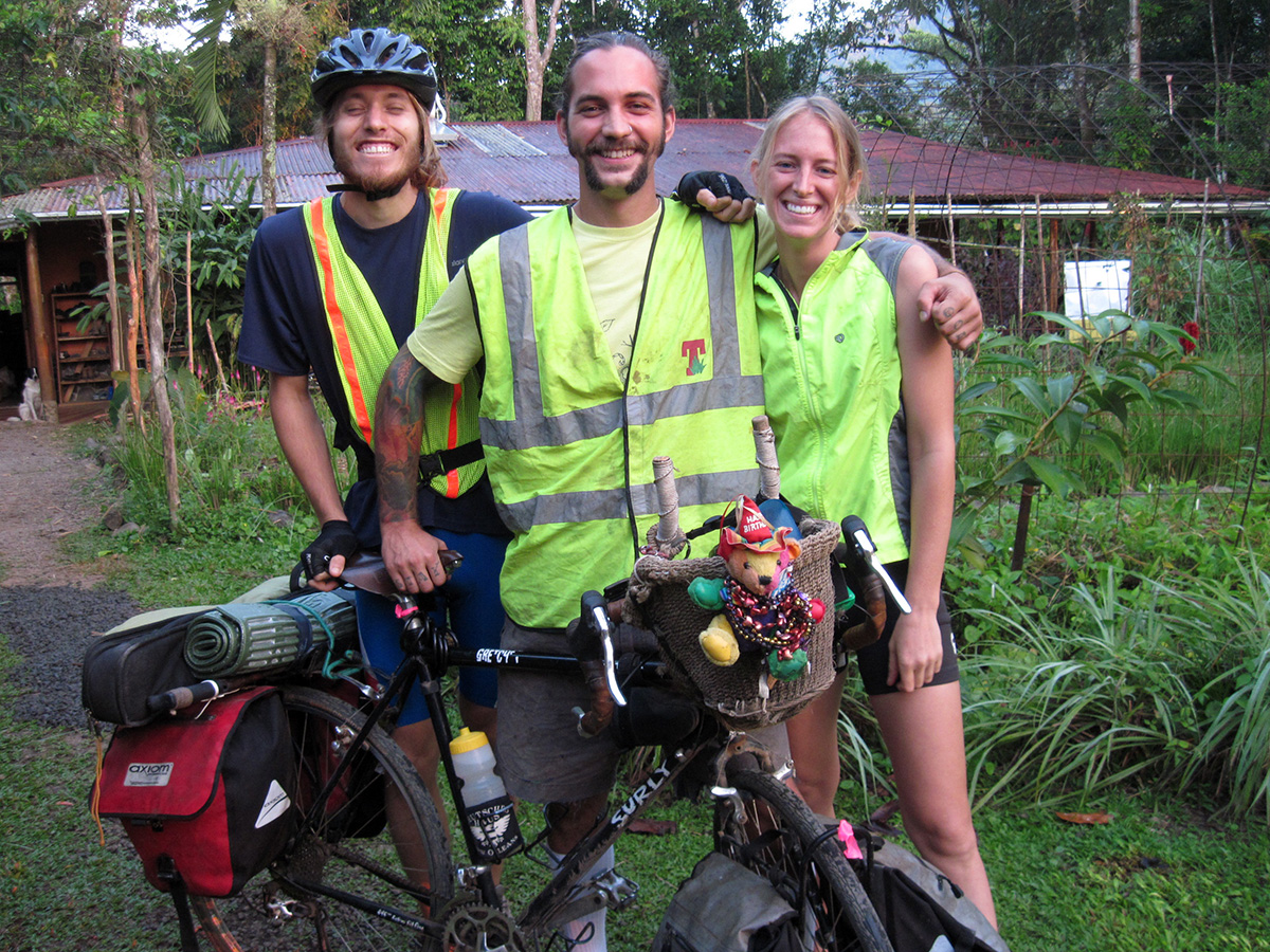 Laura, Scott, and Simon stand for a group photo on the side of the road in Costa Rica. They're all wearing neon visibility vests and have huge smiles on their faces.