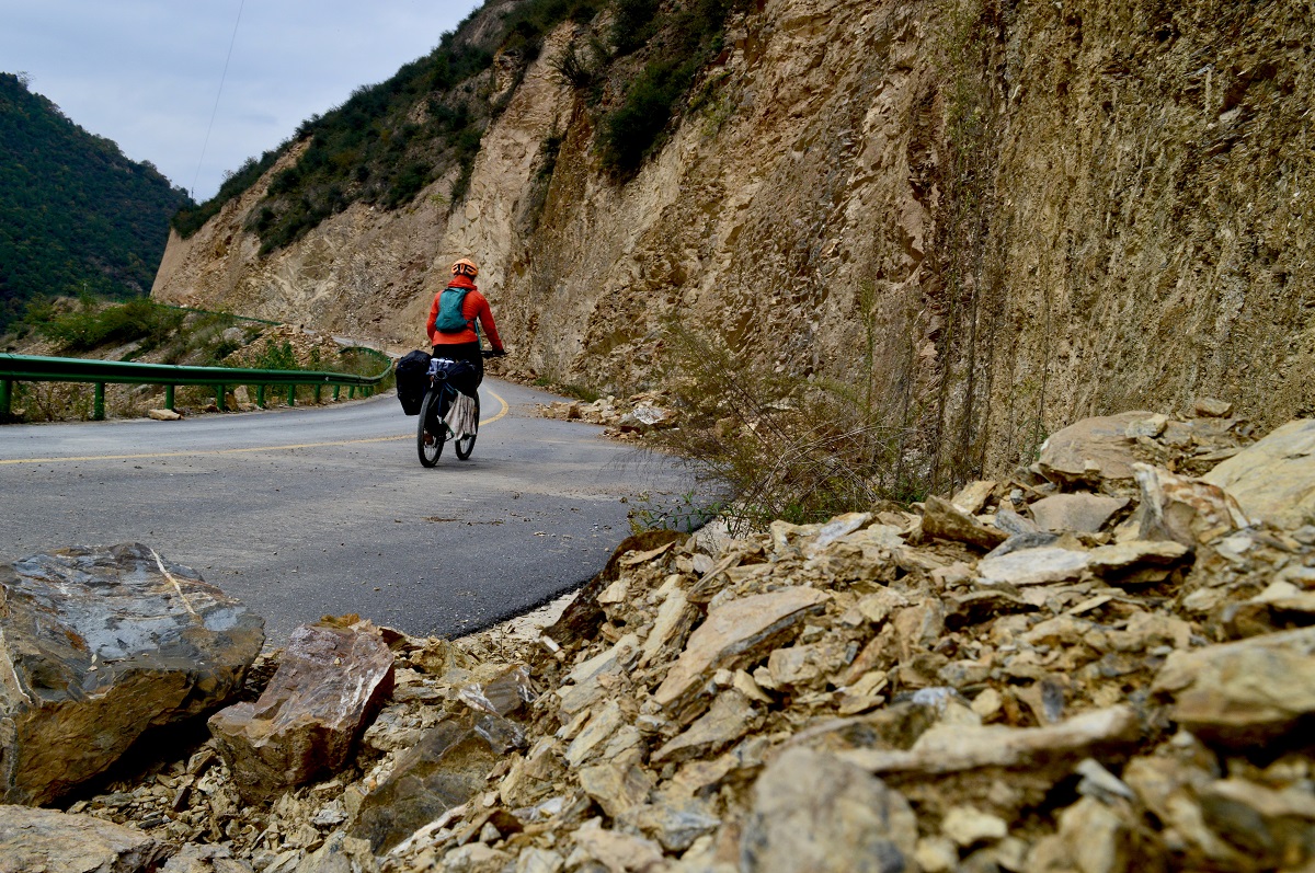 The author rides along a paved road in the mountains with fallen rocks from an earthquake at the front of the frame.