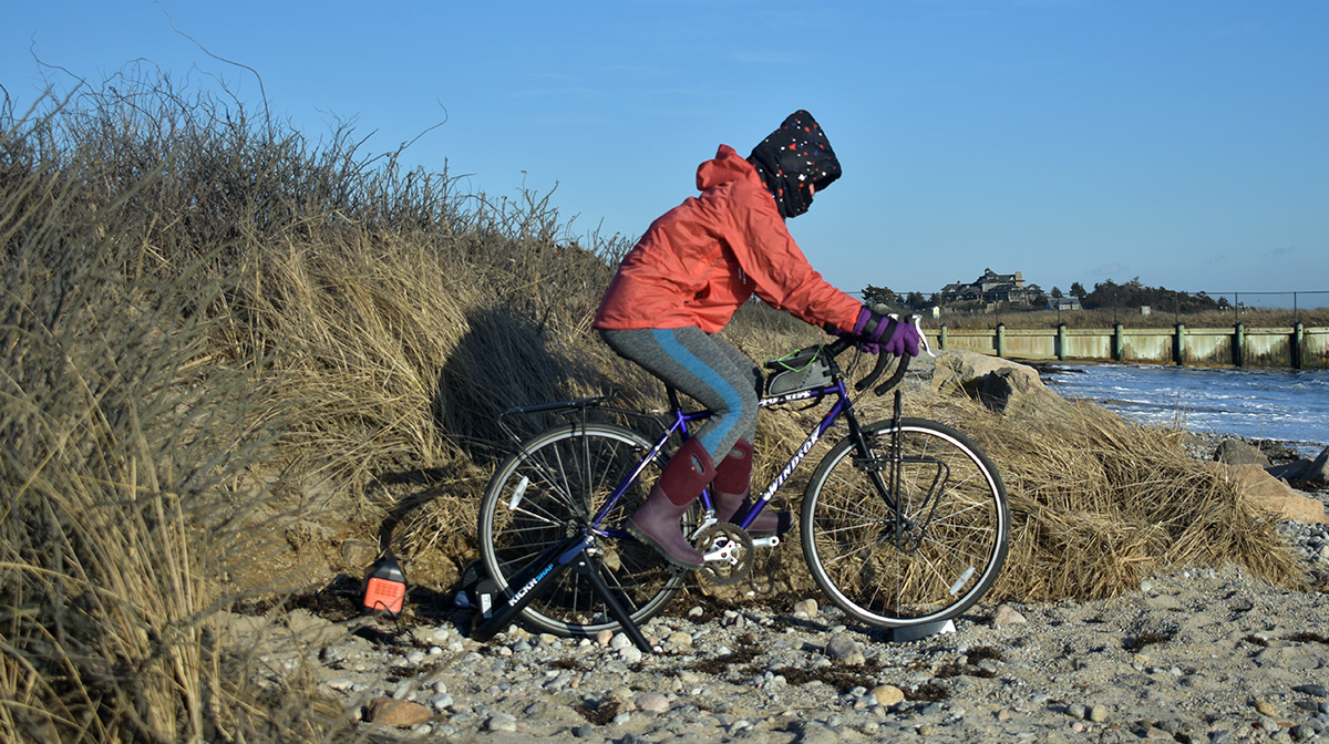 Image shows Laura riding a bike on a stationary bike stand on a sunny day at the beach. She is wearing big boots, coats, and her head and face are wrapped in scarves.