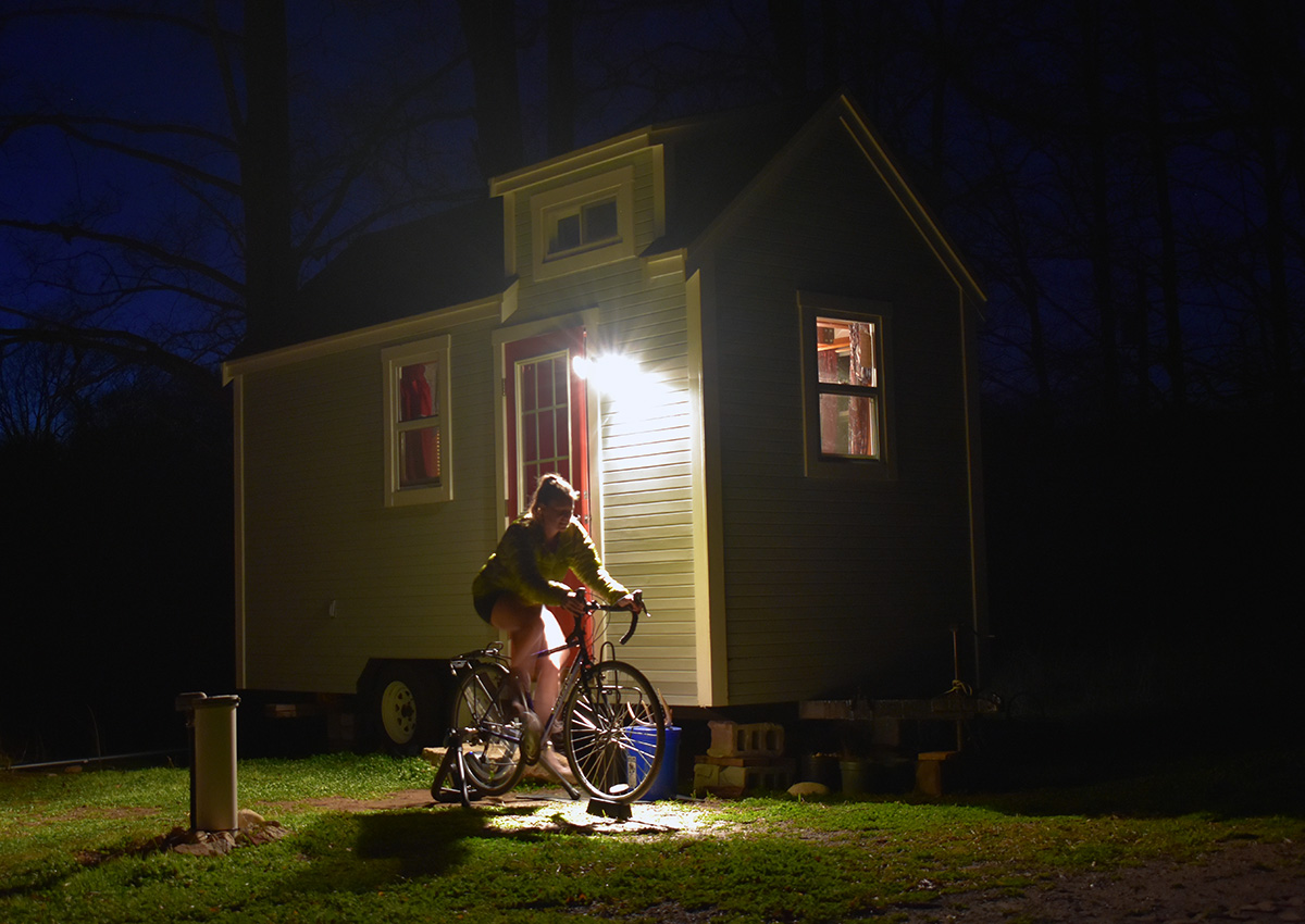 Image shows Laura riding a bike on a stationary bike stand in the dark. The light on the front of her tiny house illuminates her. The grass is noticeably green and she rides in shorts.