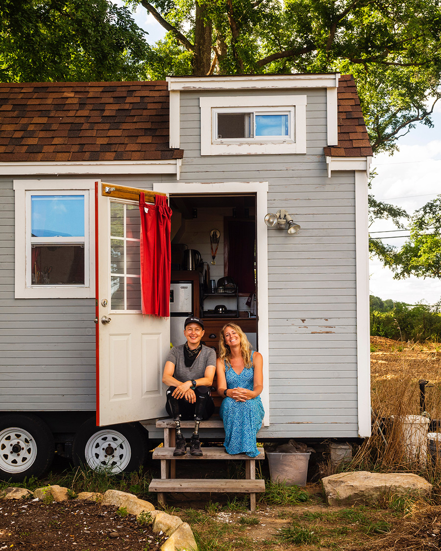 Laura and Kaisa, both white women, sit on the steps of Laura's blue tiny house on wheels among a verdant rural scene.