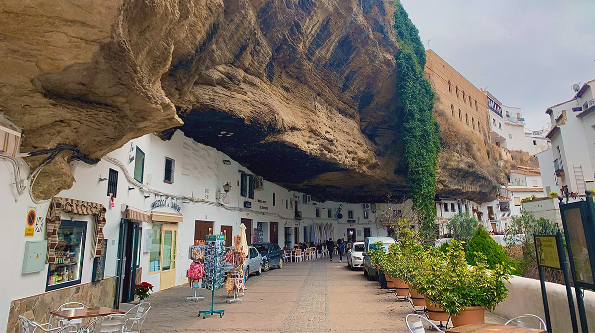 A small street makes its way along the base of a cliff under which small white houses and stores are built into the face of the cliff.