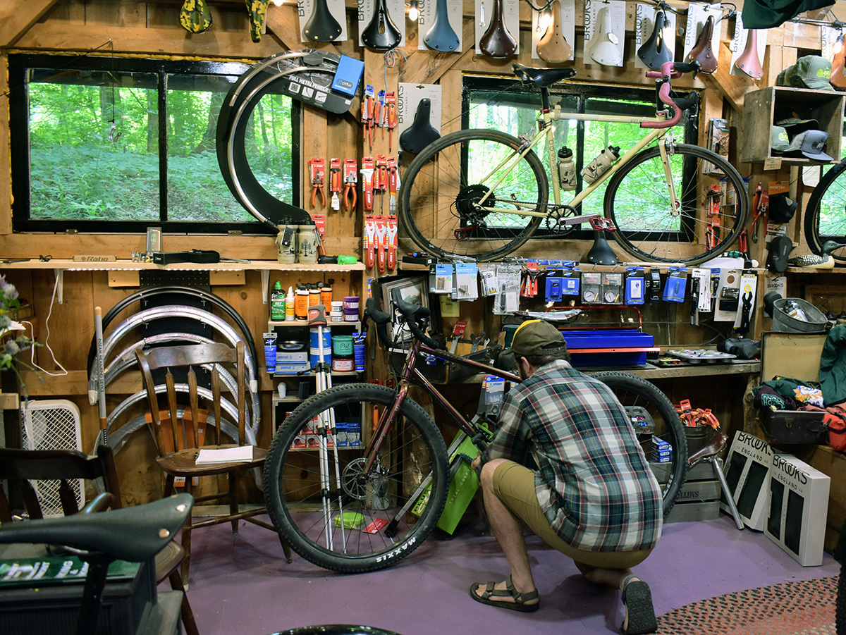 A man squats next to Laura purple bike in a log-cabin-esque bike shop that is filled to the brim with parts and saddles.