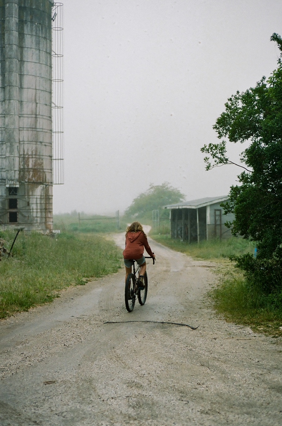 Laura rides her bike away from the camera on a dirt road, her blond hair flapping behind her. An old grain elevator and farm equipment sits to the side.