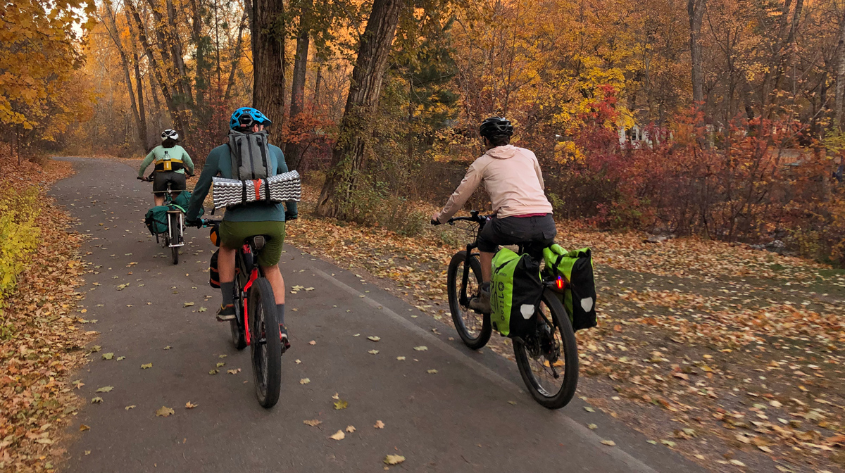 Three staff members of Adventure Cycling ride down a bike path through a park full of orange autumnal trees.