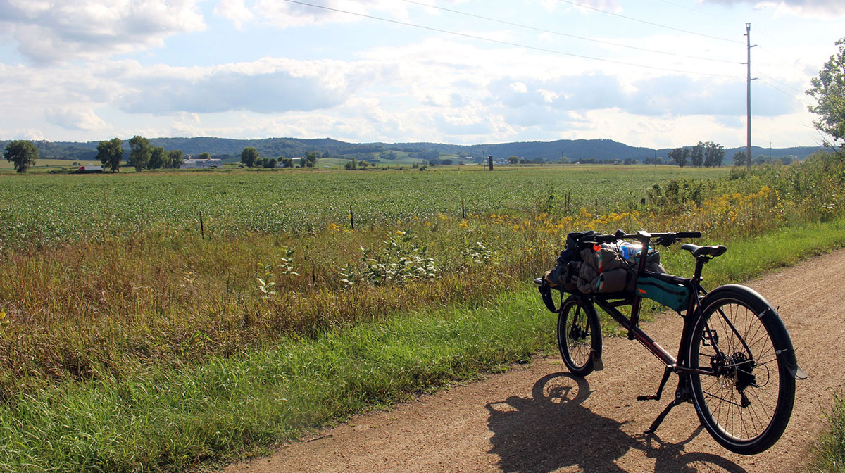A loaded cargo bike sits on a dirt rail trail overlooking trees and farmland for many miles.