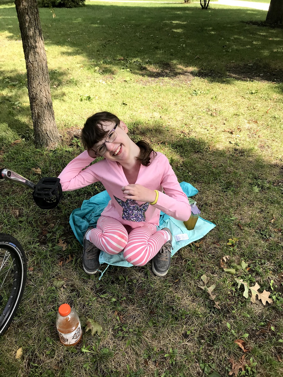 Lilia, a white girl with brown hair wearing a pink outfit, squats on the grass next to her bike and smiles into the camera