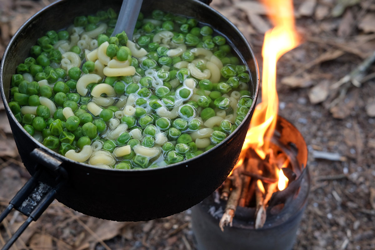 Laura holds a pot of pasta and peas over a wood fire