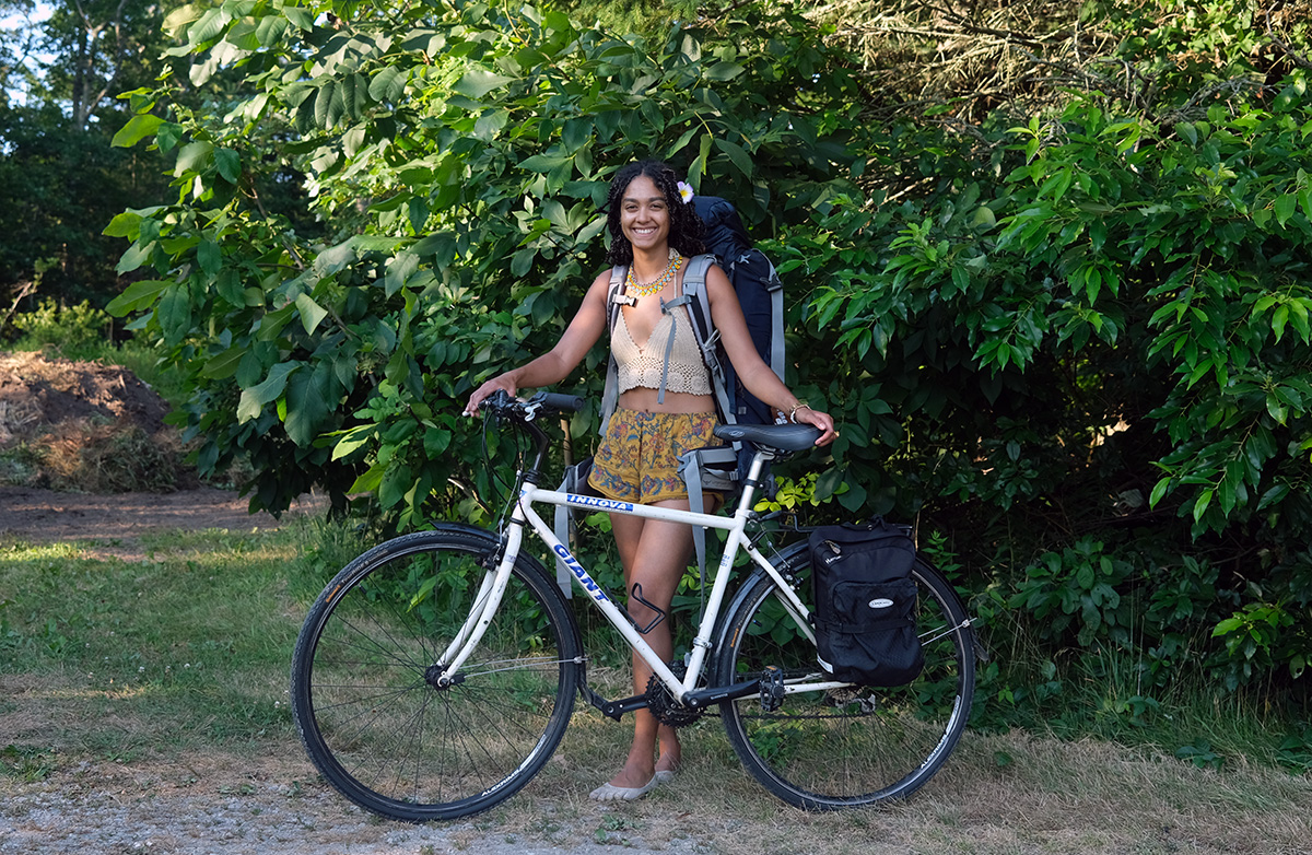 Aimee, who is female and black, poses with her borrowed bicycle under the shading foliage of a tree.