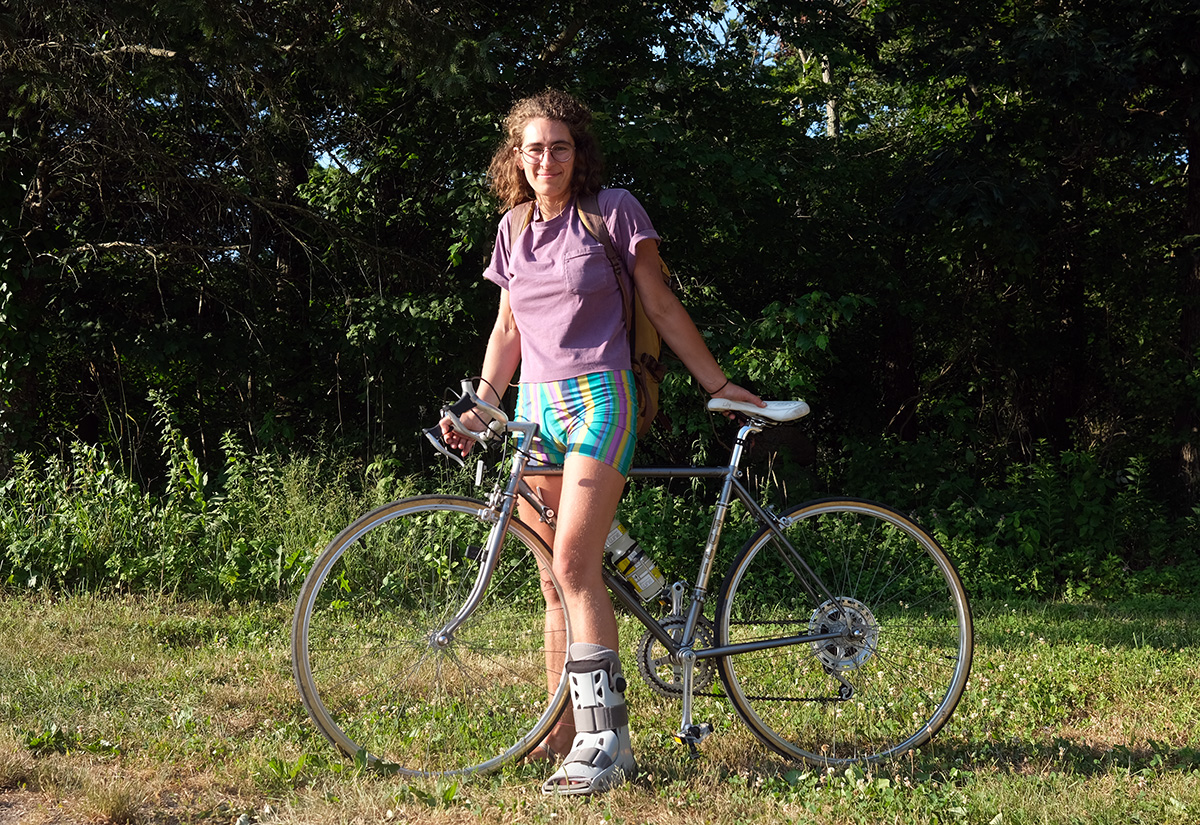 Anni, who is a white woman, poses astride her old steel road bike and wears a boot cast on her left foot