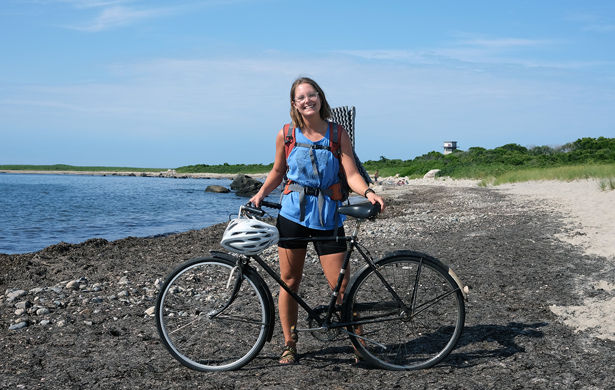 Madlyn, who is a white woman, stands on the ocean shore just inside the reach of the surf with her bicycle.