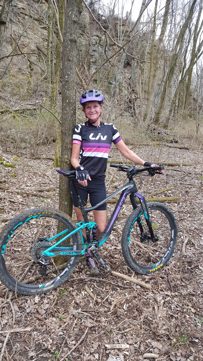 Grace Ragland is a slender older woman with blond but graying long hair. She stands with her Liv brand mountain bike in a forest.