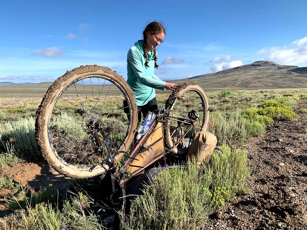 A young woman wipes thick, sticky mud off of the tires of her upturned bike in the middle of a vast and remote landscape in New Mexico