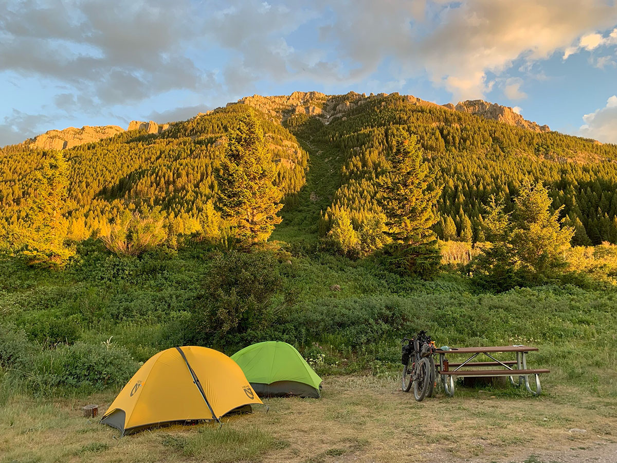Two tents, one green and one yellow, sit among a beautiful scenery of trees and the last of evening sunlight.