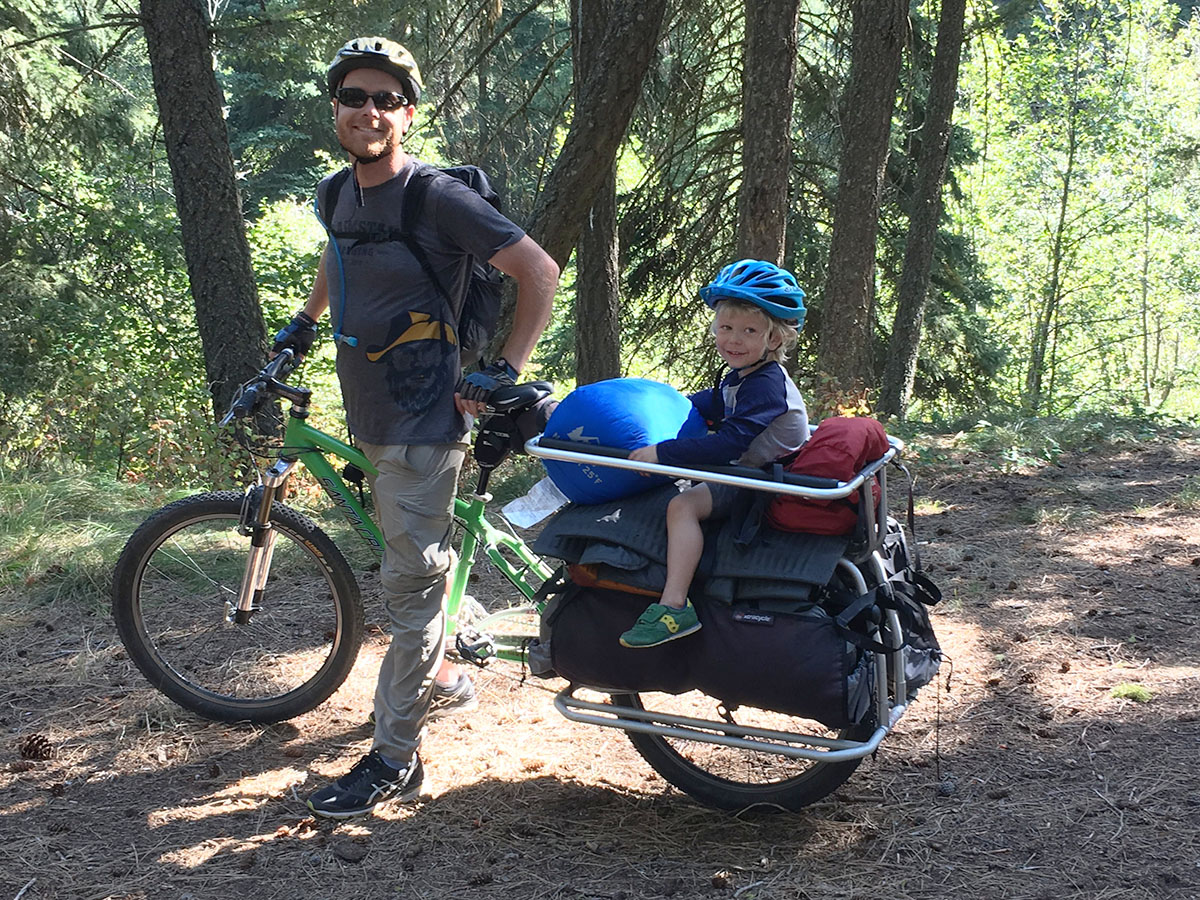 A dad stands over his upright cargo bike while his son sits on the cargo part in the back