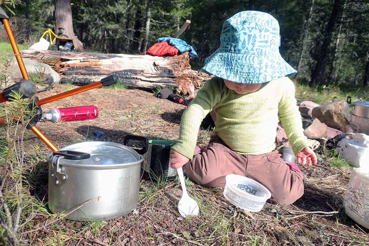 A toddler in a bucket hat digs in the dirt with a camp spoon