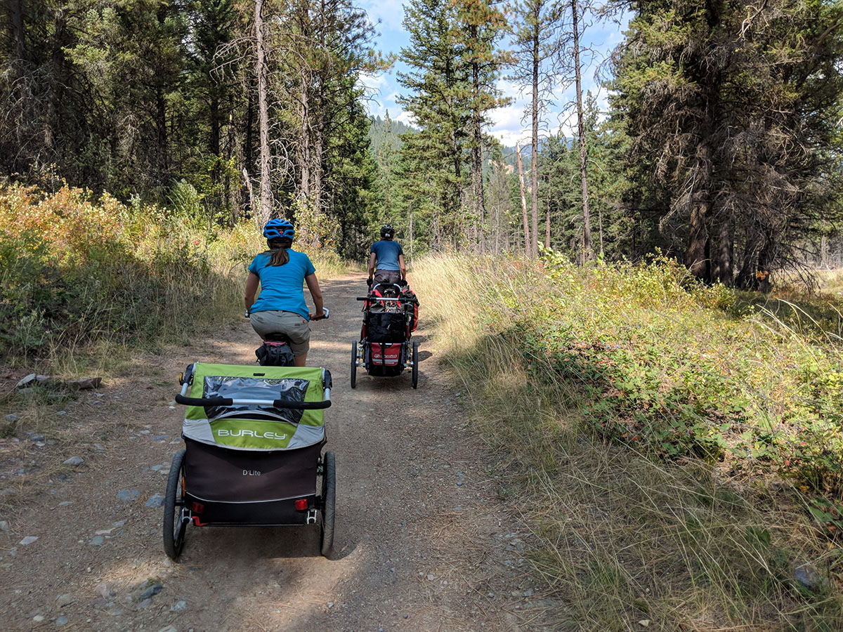 Two parents with trailers full of kids and gear ride on a wide dirt path through the forest.