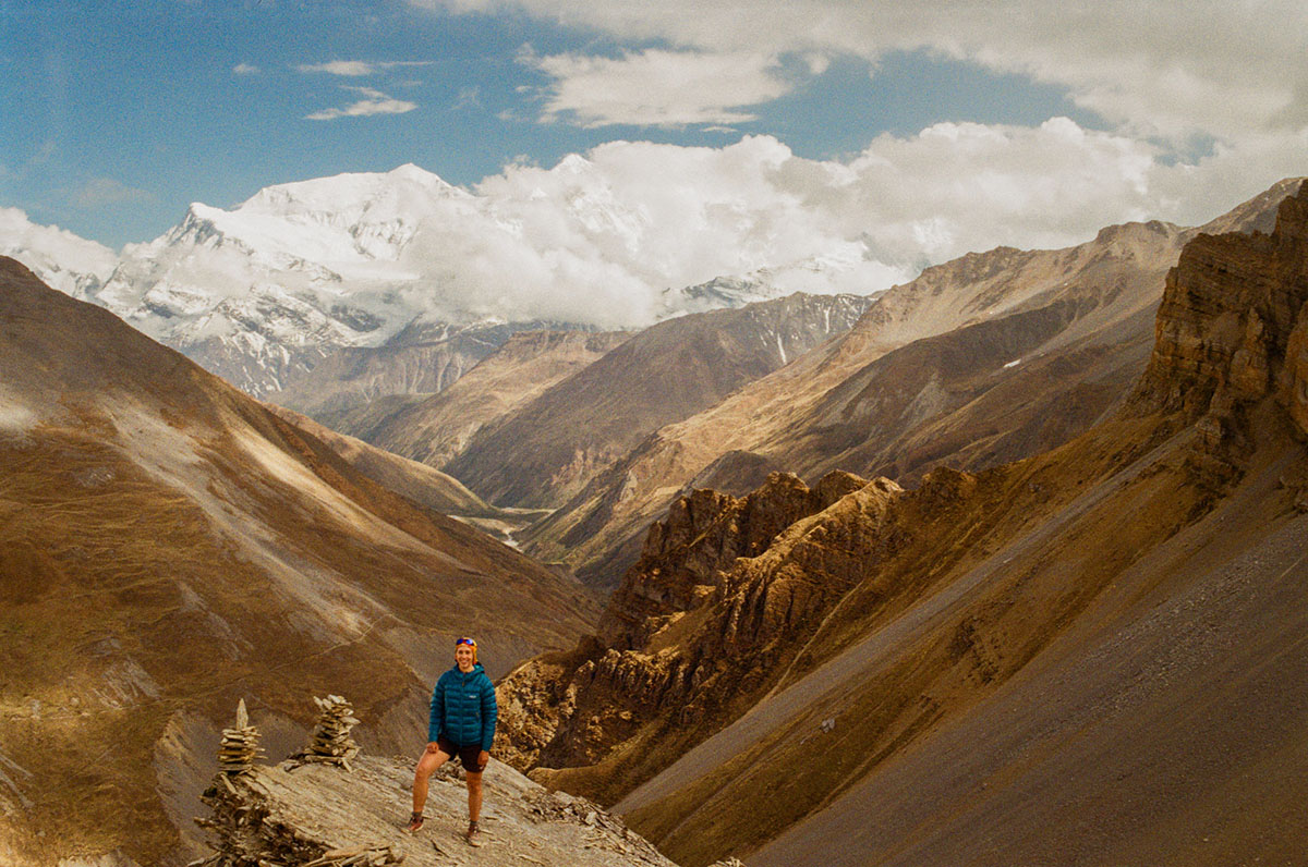 Mia, a white woman, stands on a precipice bundled in warm clothes as dramatic, snow capped peaks jut into the sky behind her. The landscape around her is rocky, brown, and devoid of vegetation.