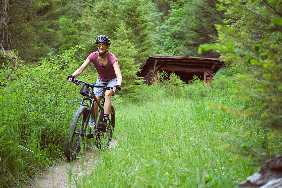 Photo shows author riding a front suspension mountain bike on a forested trail while wearing the Ride Concept shoes, gray shorts, and a maroon shirt. Author is a white female with brown hair and long limbs.