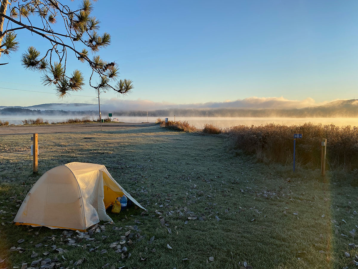 A yellow tent sits under a tree near a lake. Morning mist hovers above the lake surface and frost lies lightly on the top of the grass.