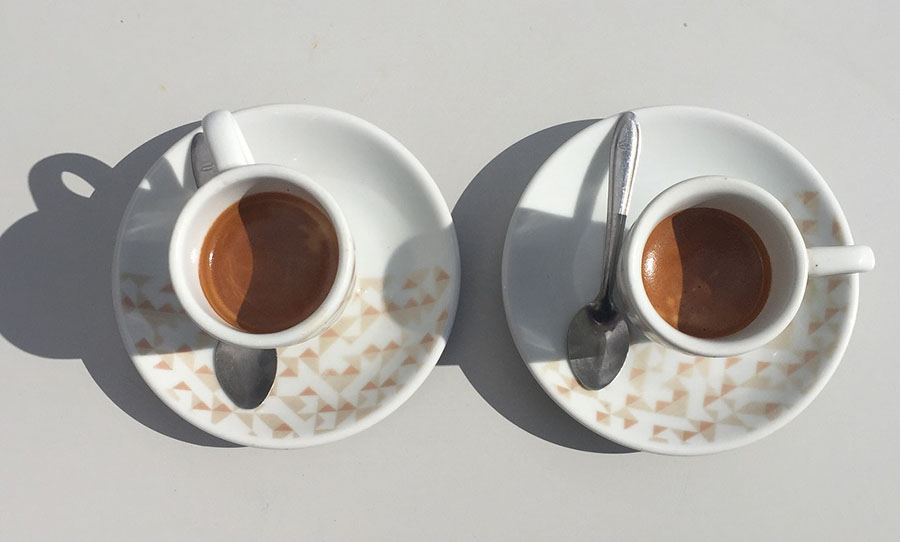 Two cups of espresso sit on small matching plates on a white outdoor table, the cups casting long shadows.