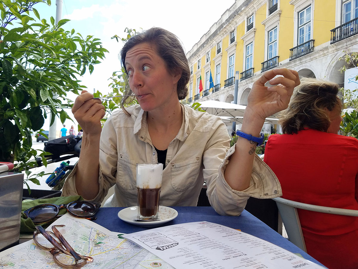 Hollie sits at a small outdoor dining table in a Portuguese town, sipping latte froth from a spoon. 