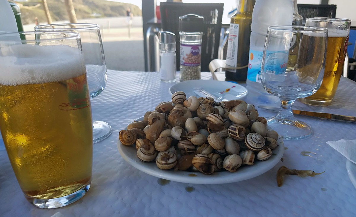 A bowl of snails in the shell sits on an outdoor table with two yellow beers.