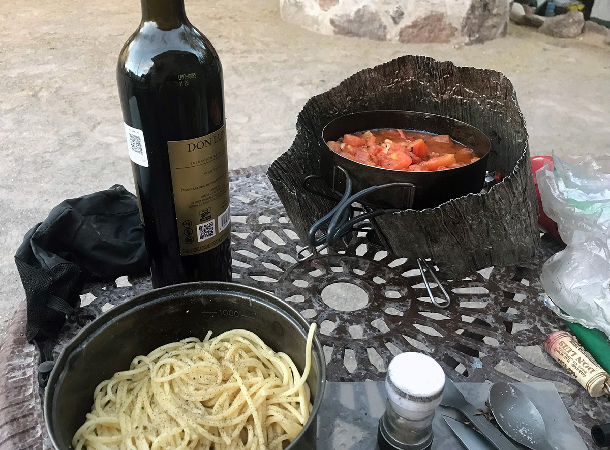 A bottle of red wine, a pot of peppered pasta, and a pot of fresh tomato sauce sit on an outdoor table, ready to be devoured.