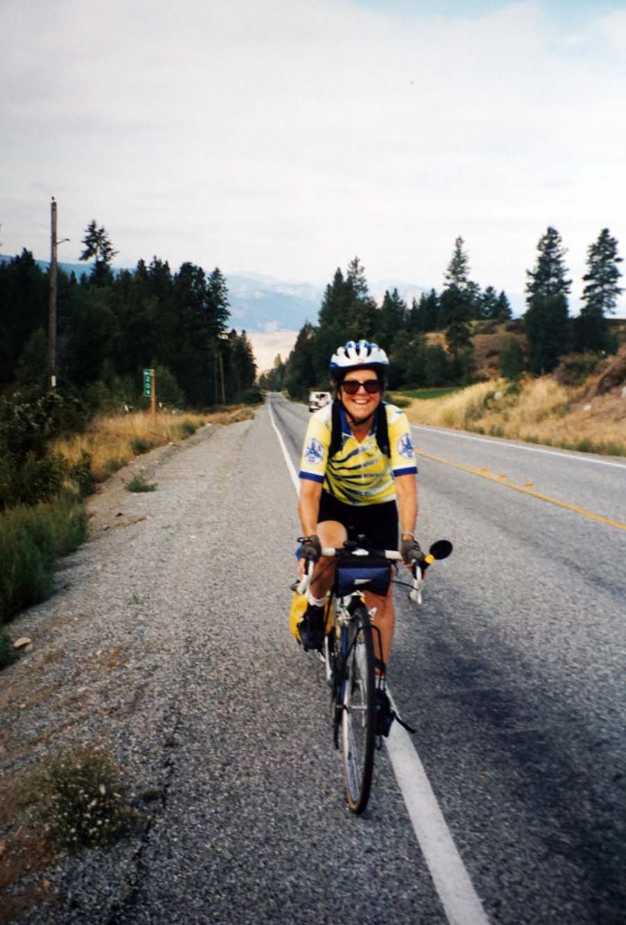 Aunt Liz grins while riding on the shoulder of a rural highway. There are trees along the roadway and mountains in the background.