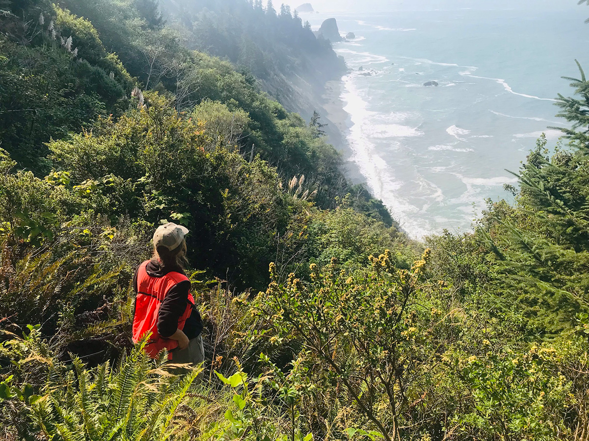 A woman stands in the bushes near some rocky cliffs overlooking the Pacific Ocean.