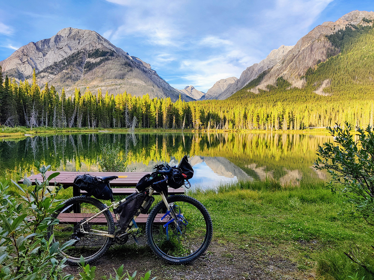 Alissa's loaded bike rests upright against a picnic table next to a mountain lake. The lake is surrounded by pine trees with a backdrop of sharp, rocky mountains that are lit by the setting sun.