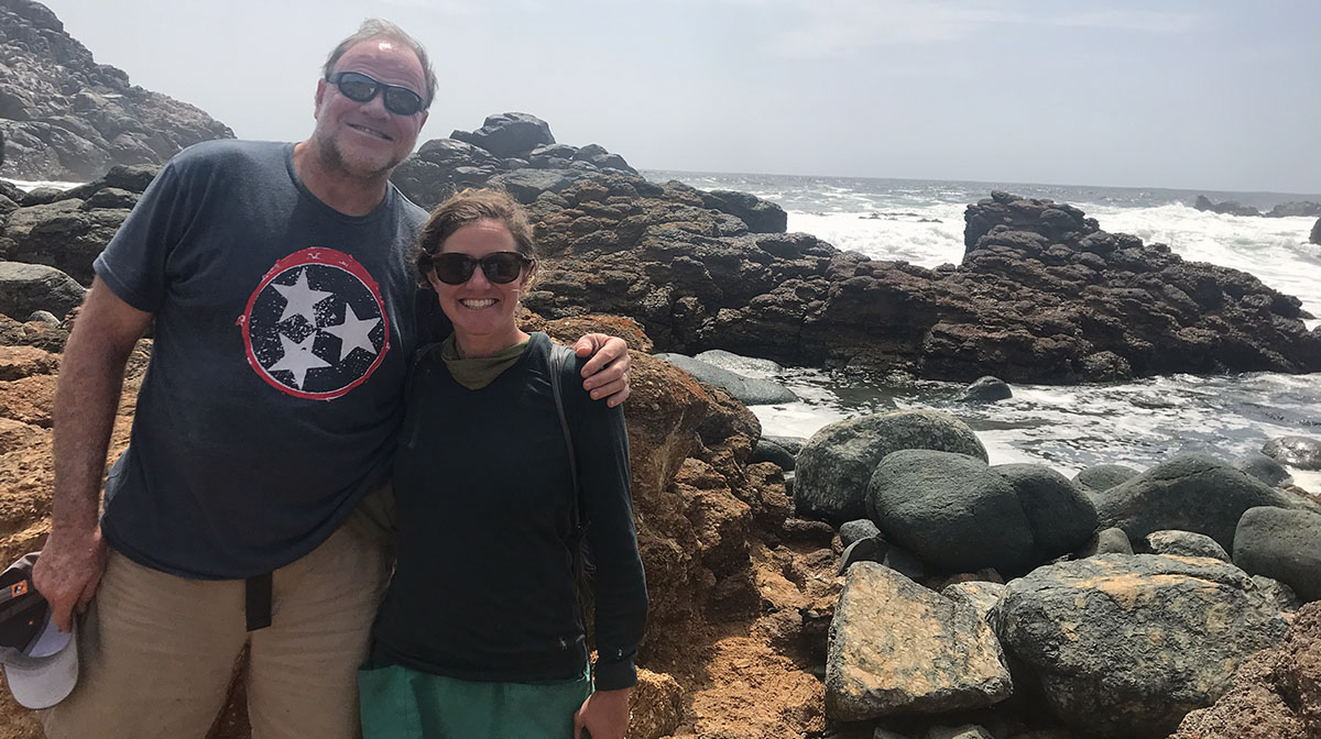 Hollie and her dad stand next to the ocean on a brown, rocky outcropping. Their hair is blowing in the wind.