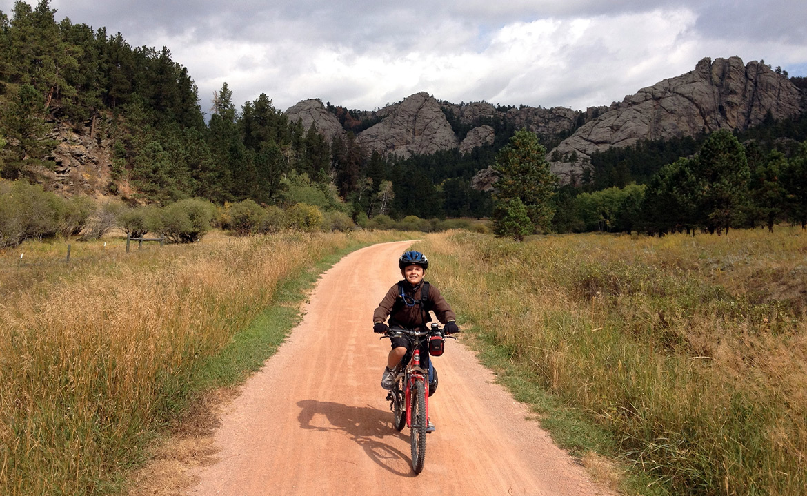 A little boy rides his bike on a dirt bike trail with rocky, rolling hills with few trees, mostly shrubs, in the background. He's smiling at the camera.
