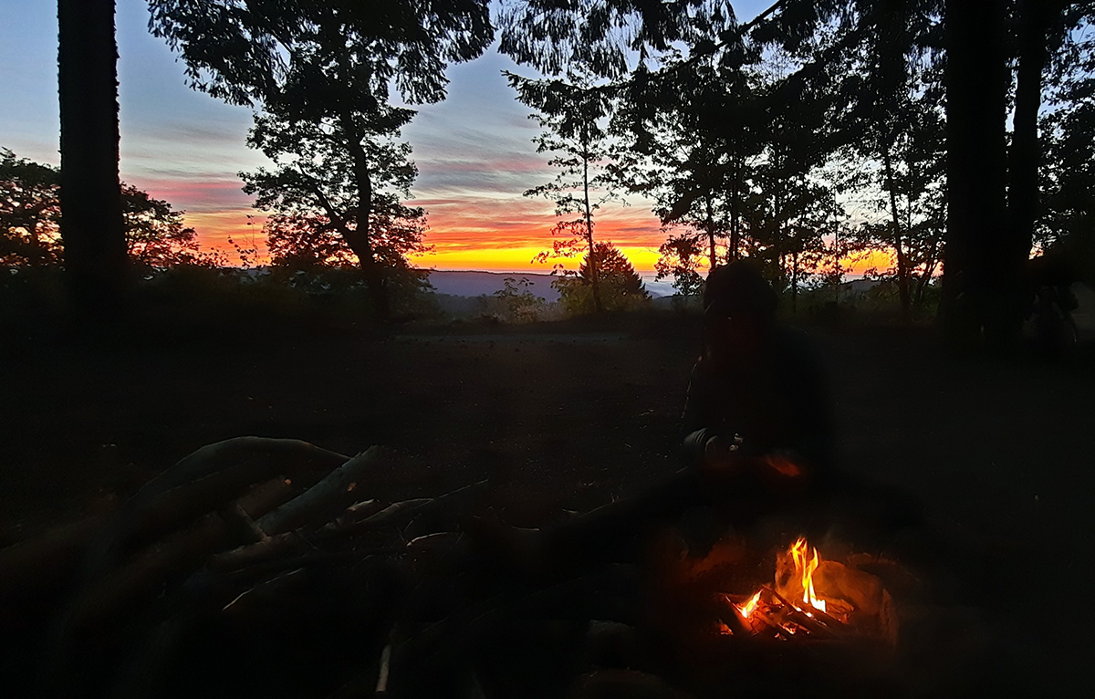 The view of a sunset from atop a forested mountain with a campfire in the foreground.