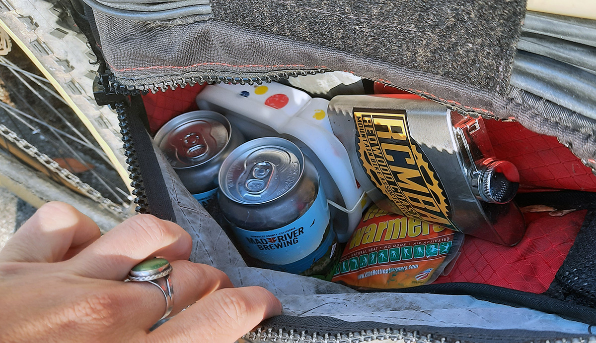 A photo of the contents of a frame pack: two beers, one sandwich, a replacement tube, a flask of whiskey, and emergency handwarmers.
