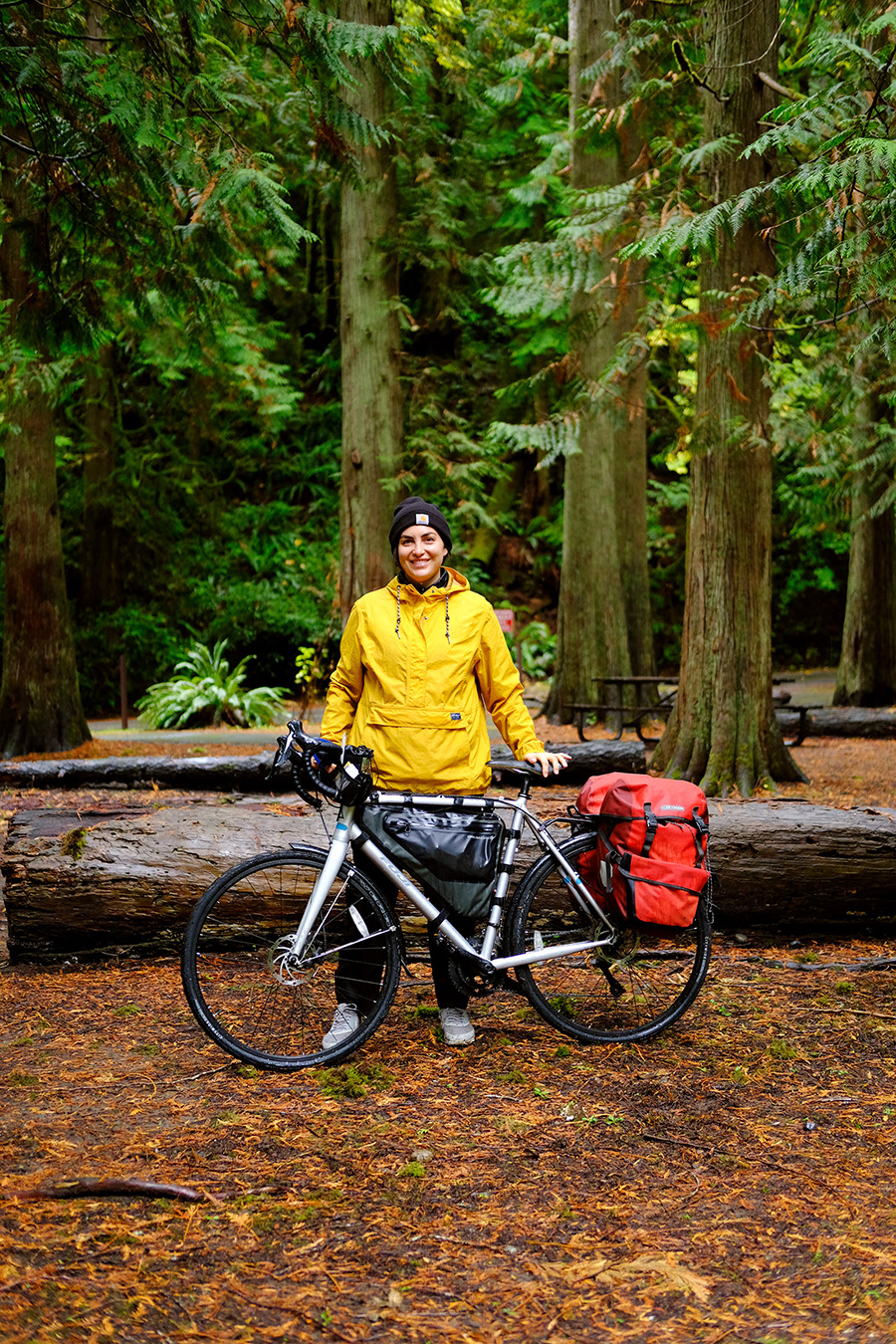 Roxy wears a bright yellow rain jacket as she stands among tall cedar trees with her loaded gravel bike