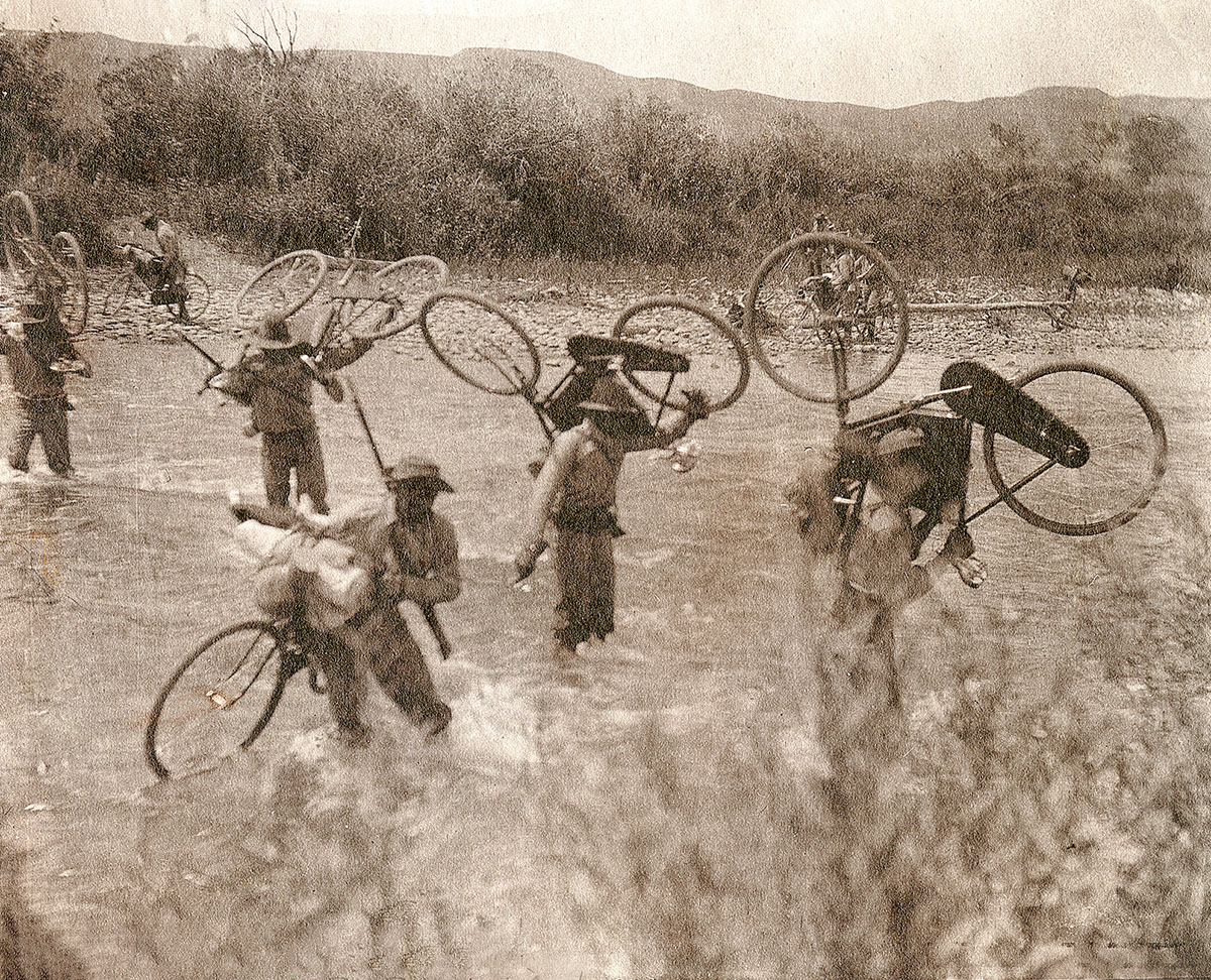 The soldiers wade across a river, their bikes being carried on their backs.