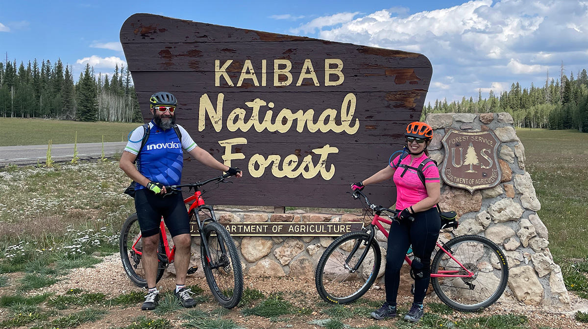 Two people stand in front of a National Forest sign reading Kaibab National Forest.